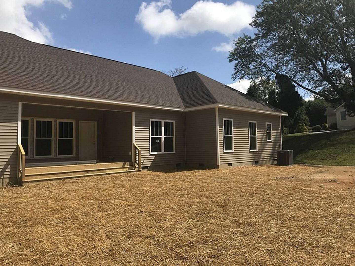 Two-story house with gray siding, covered front porch, white trim, large windows, manicured lawn, mature tree, and blue sky with scattered clouds