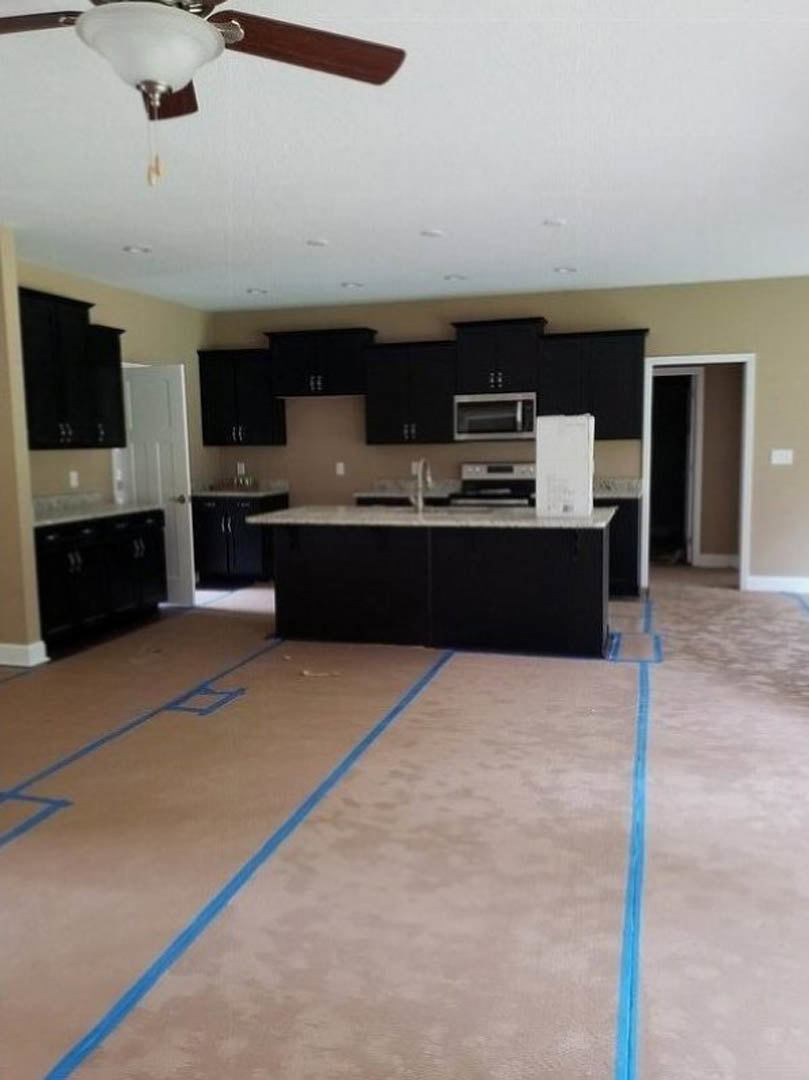 Kitchen with matte black cabinets, white countertops, blue painter’s tape on the floor, ceiling fan with light fixture, and a window in the background.
