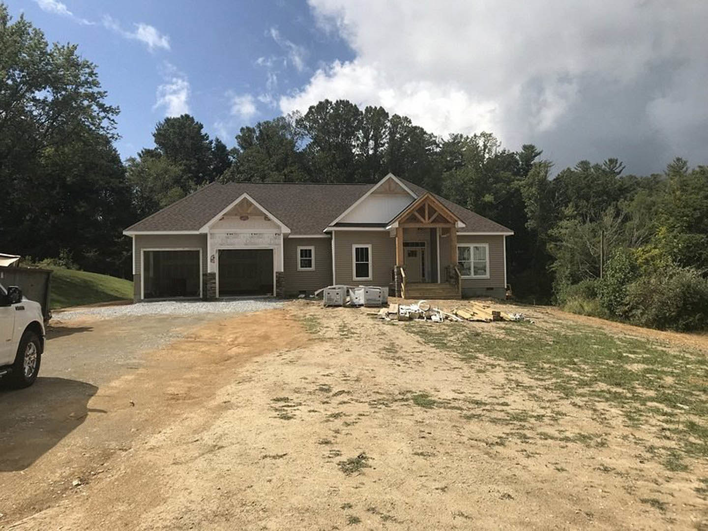 Modern house with attached garage, surrounded by dirt yard and mature trees, white pickup truck parked in driveway, blue sky overhead