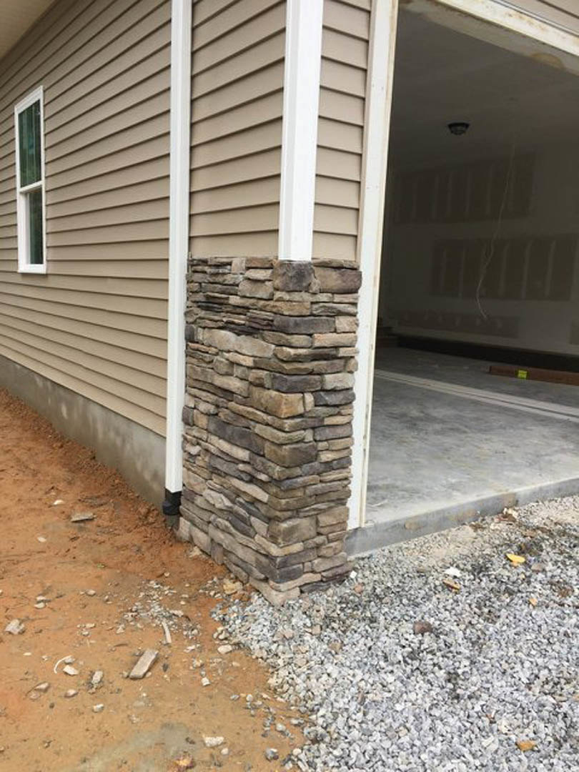 Stone pillar with stacked stone finish beside white stucco wall, white-framed window, and pile of rocks at base on residential home exterior