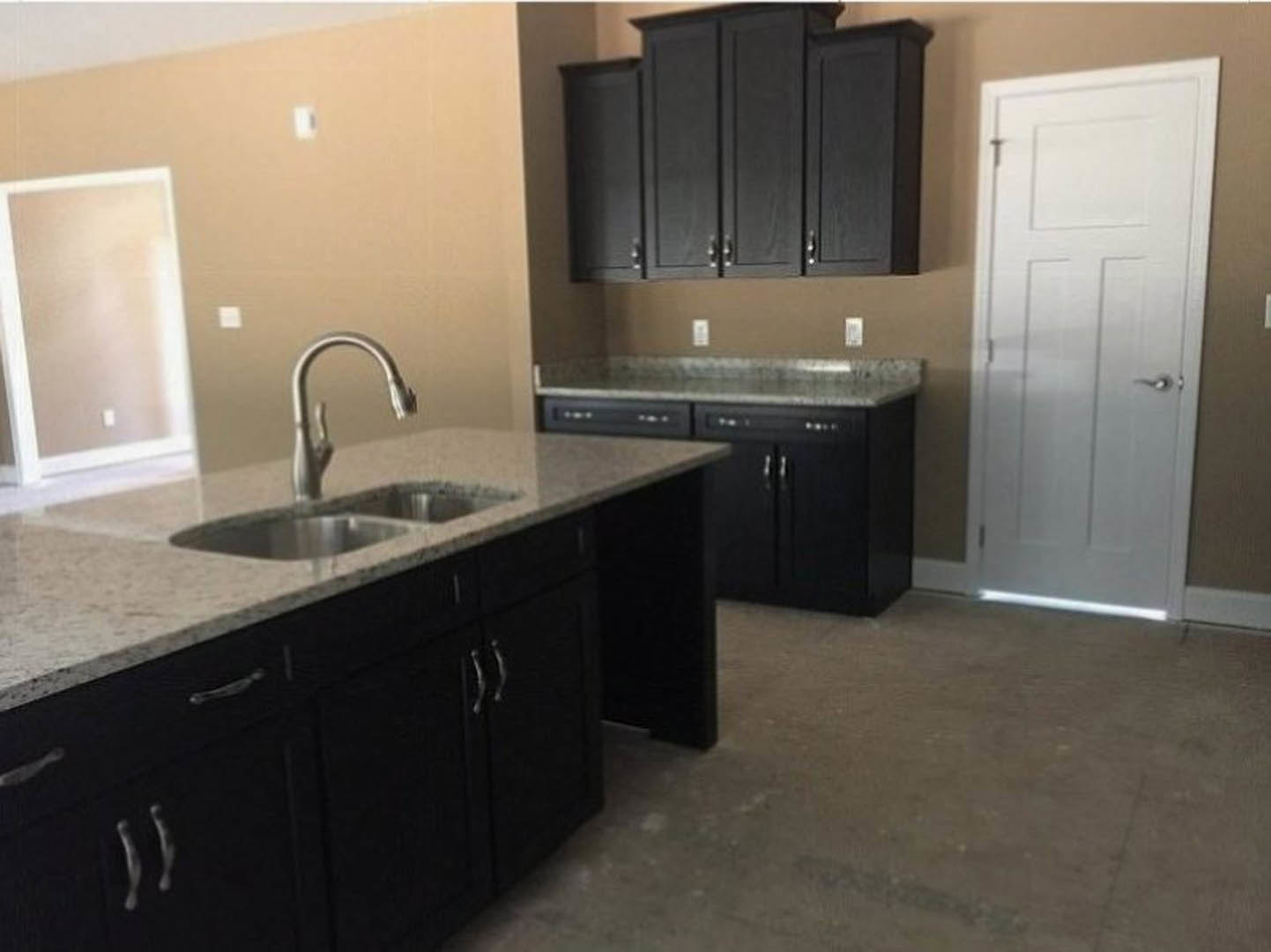 Black kitchen cabinets with granite countertops, stainless steel faucet and sink, white door with silver handle, white wall featuring a light switch, tile flooring