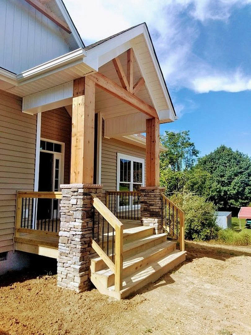 Front porch with wooden stairs, stone pillar, metal railing, and large window, surrounded by trees and horizontal siding