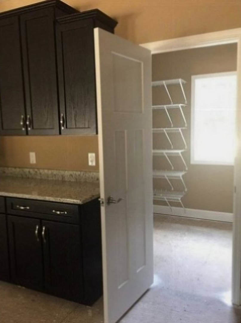 Modern kitchen with white cabinetry, open door revealing pantry shelves, black-edged countertop, wire rack near window, and light wood flooring