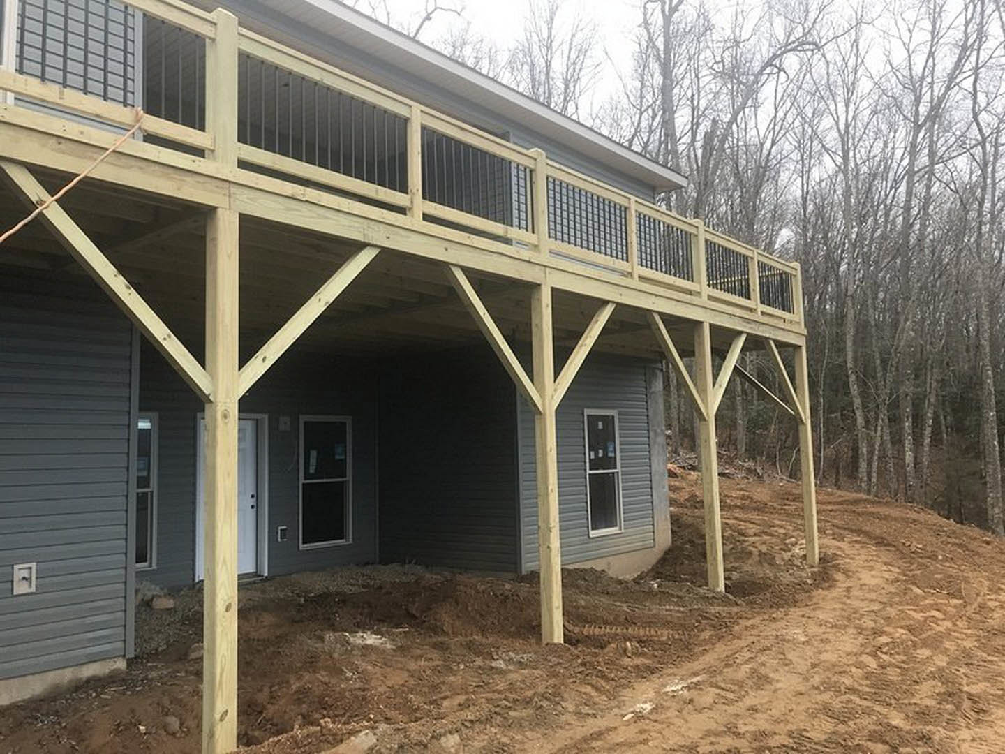 Two-story home with gray siding, white-framed windows, elevated wooden deck, covered porch, dirt path leading to entrance, and mature trees in the background