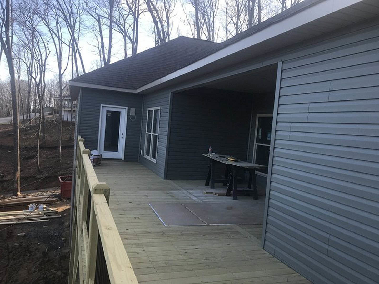 Wooden deck attached to a house with light siding, outdoor table and chairs, large windows, and surrounding trees
