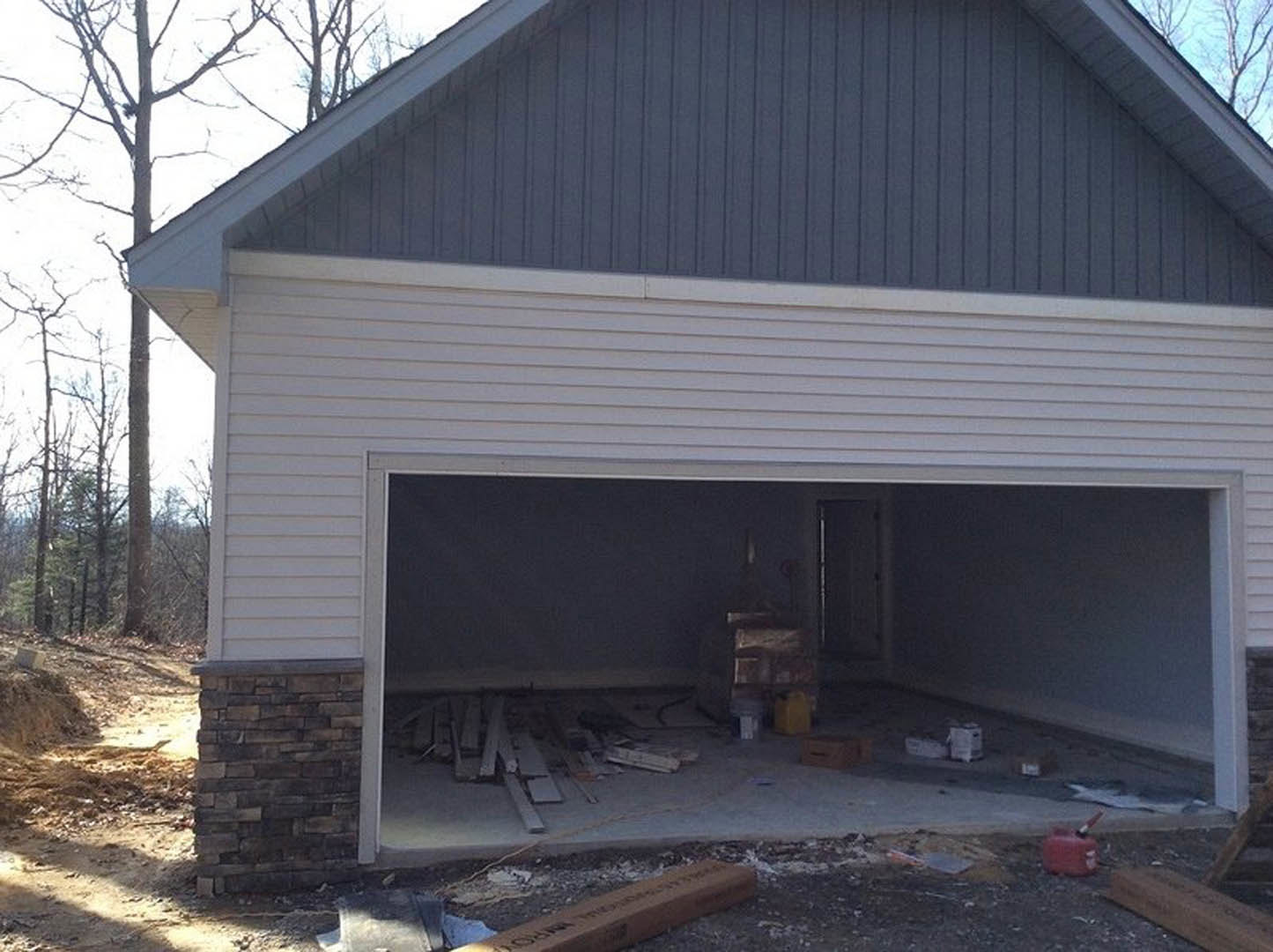 Partially built garage with exposed wooden framing, brick wall with white trim, construction materials scattered on concrete floor, unfinished roof structure, black rectangular