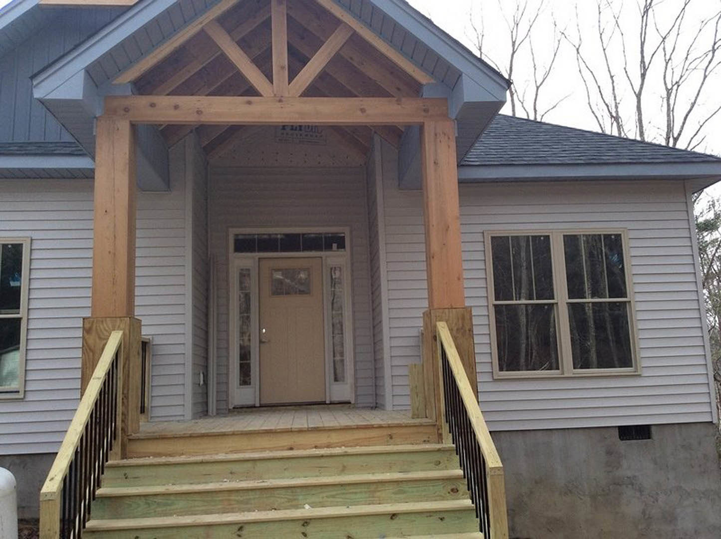 Front porch with wooden stairs, white siding, large windows, and dark front door; trees visible through windows; exposed wooden roof trusses above entry.