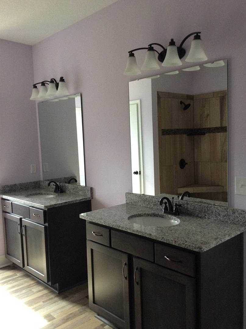 White bathroom with rectangular sink, chrome faucet, stone countertop, wood cabinetry, large framed mirror, and light gray tile walls