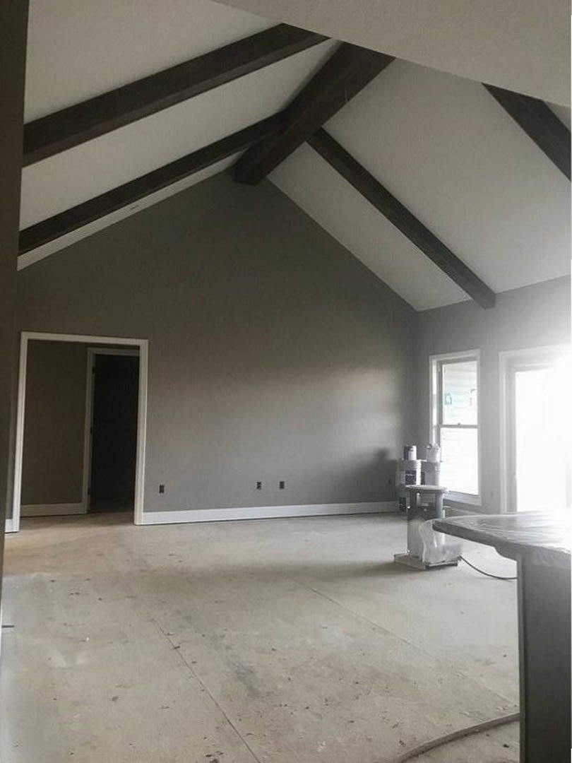 Dining area with exposed ceiling beams, white plaster walls, rectangular wooden table, black door with white frame, and large window providing daylight