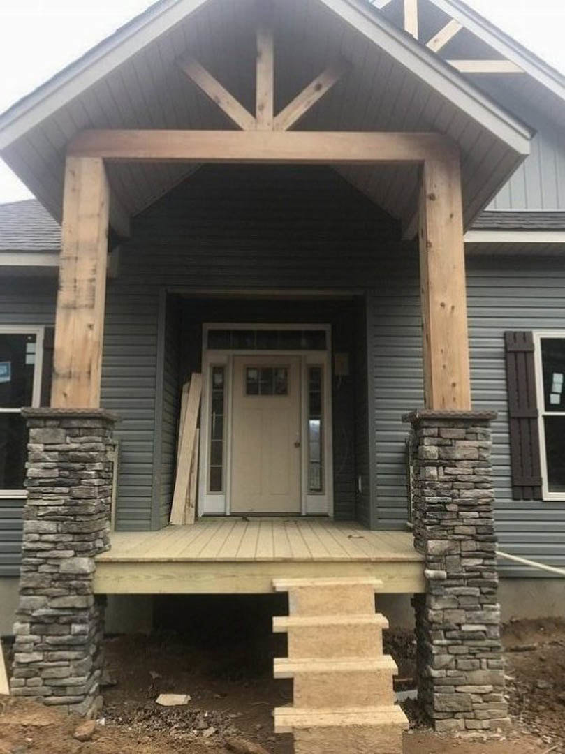 Stone staircase leads to covered porch with wooden railing, white door with glass window, stone pillar, and light-colored siding.