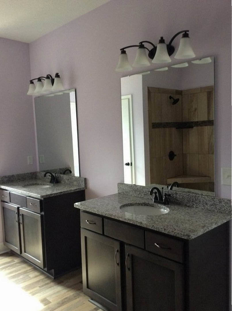 White bathroom with rectangular sink, wall-mounted mirror, light gray tile backsplash, wood cabinetry, chrome faucet, and stone countertop.