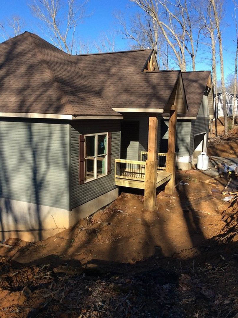 Framed house under construction with exposed wooden porch, unfinished dirt yard, white window trim, and trees in the background