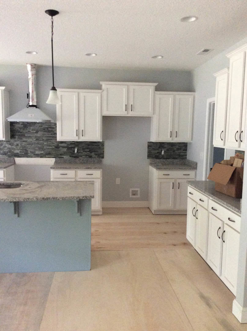 White kitchen cabinets with granite countertops, stainless steel sink, and modern appliances; black pole on white surface, cardboard box on floor, white pipe along wall.