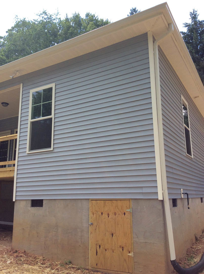 White siding house with covered front porch, wooden entry door, large window, gable roof, and landscaped yard with trees.