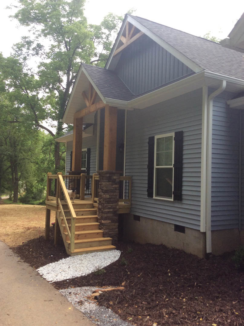 Two-story home with light siding, brick pillar, white-framed windows, wooden porch and stairs, white gravel landscaping, dog standing on deck