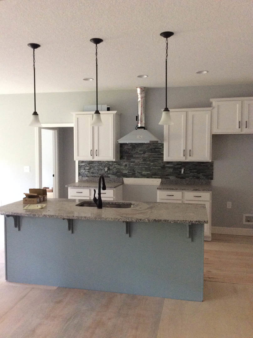 White kitchen cabinets and island with light stone countertop, built-in sink, black metal fixtures, pendant ceiling light, and wood flooring.
