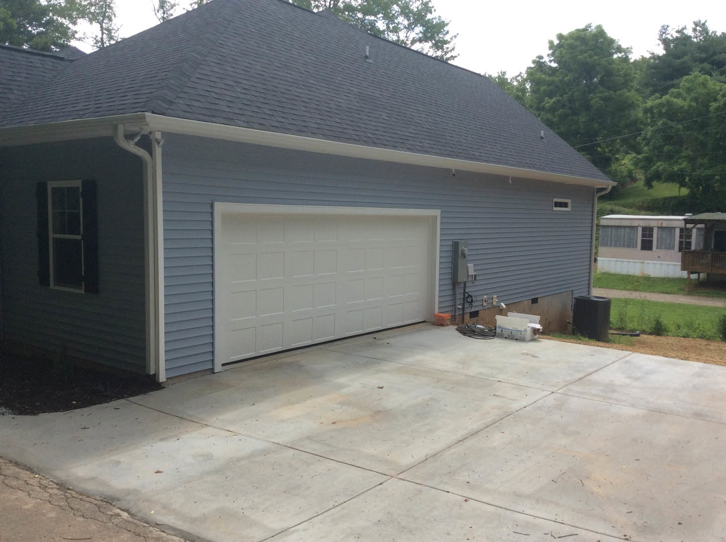 White garage door with square panels, concrete driveway, light-colored siding, small window with white frame, tree and sky in background