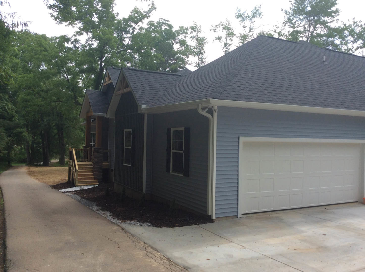 Two-story home with blue roof, white siding, attached garage with white door, paved driveway, white-framed windows, wooden stairs leading to front entrance, surrounded by trees and