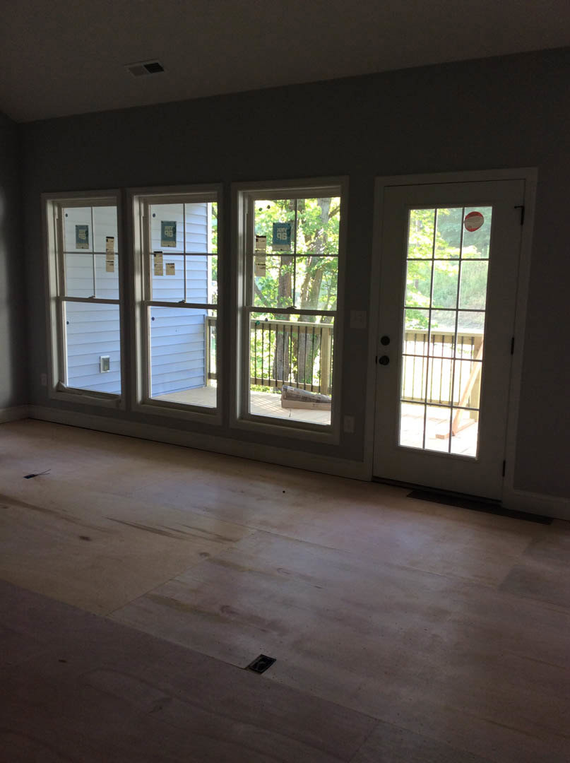 Open living area featuring large windows, glass-paneled door leading to wooden deck, light wood flooring, and views of trees outside.