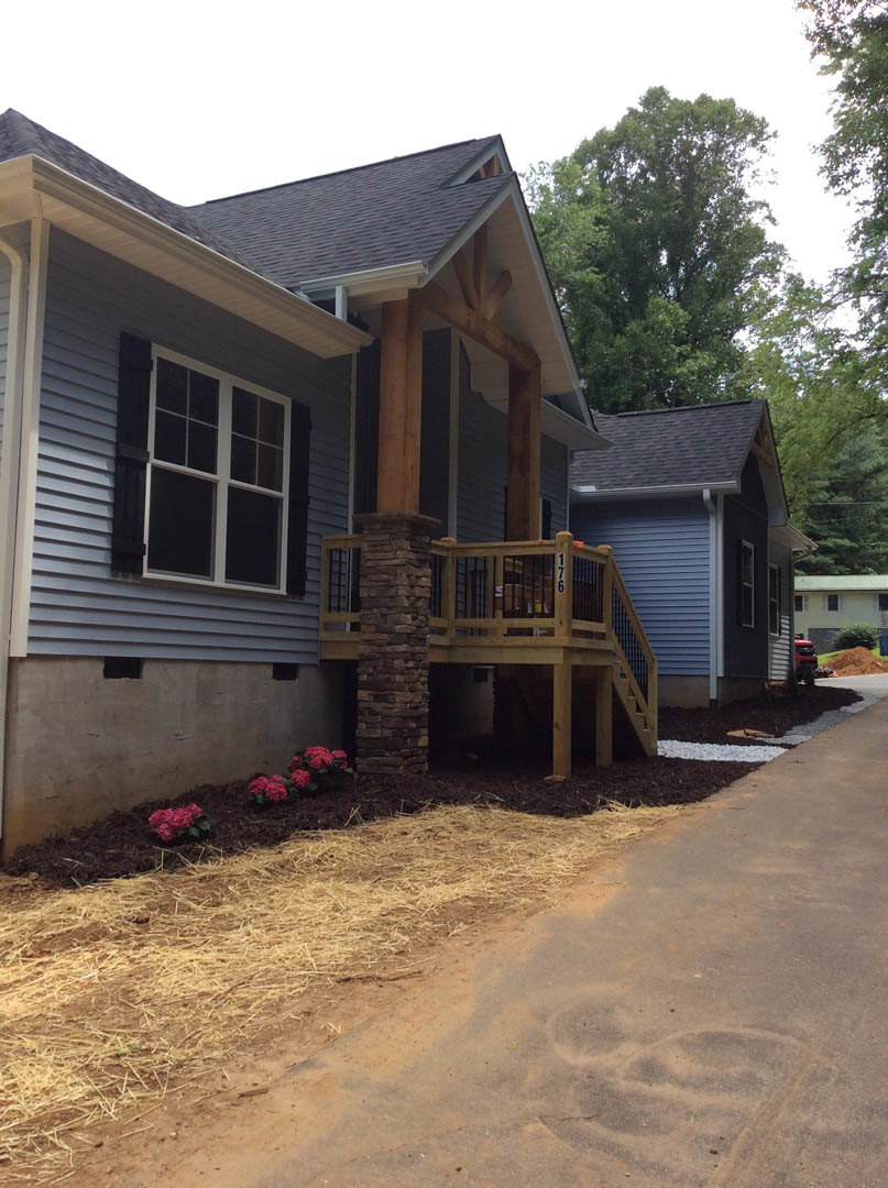 Stone porch pillar supporting wooden deck with railing, adjacent window framed in white trim, dirt path lined with straw, garden bed featuring blooming flowers, brown and black