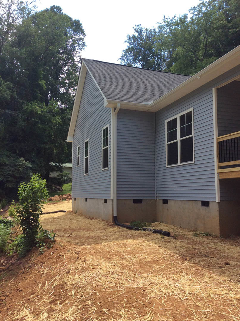 White siding house with covered porch, wooden deck, large window, green-leafed tree nearby, straw mulch along foundation, black pipe on ground, shingled roof visible in background