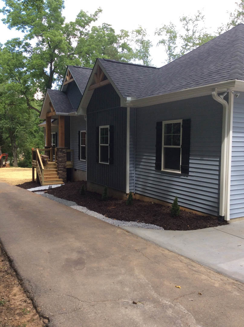 Two-story house with gray siding, white-framed windows, covered front porch, wooden stairs with railings, concrete driveway, and landscaped yard with small trees and mulch beds