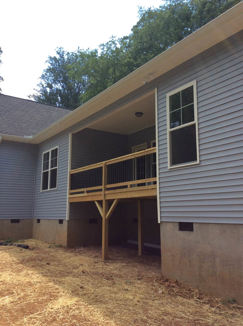 Two-story home with white-framed multi-pane windows, wooden deck, covered porch, hay-covered ground, and mature trees in the background