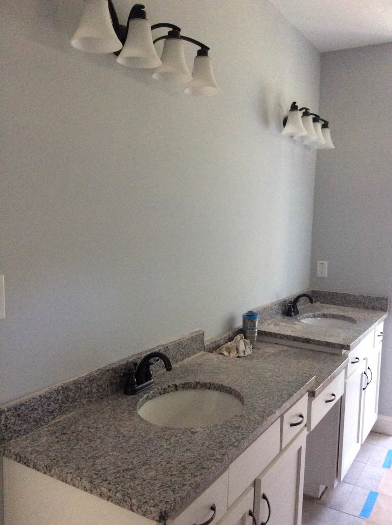 Double vanity bathroom with white sinks, matte black faucets, light gray stone countertop, white tile backsplash, wood cabinetry, and modern circular pendant lights