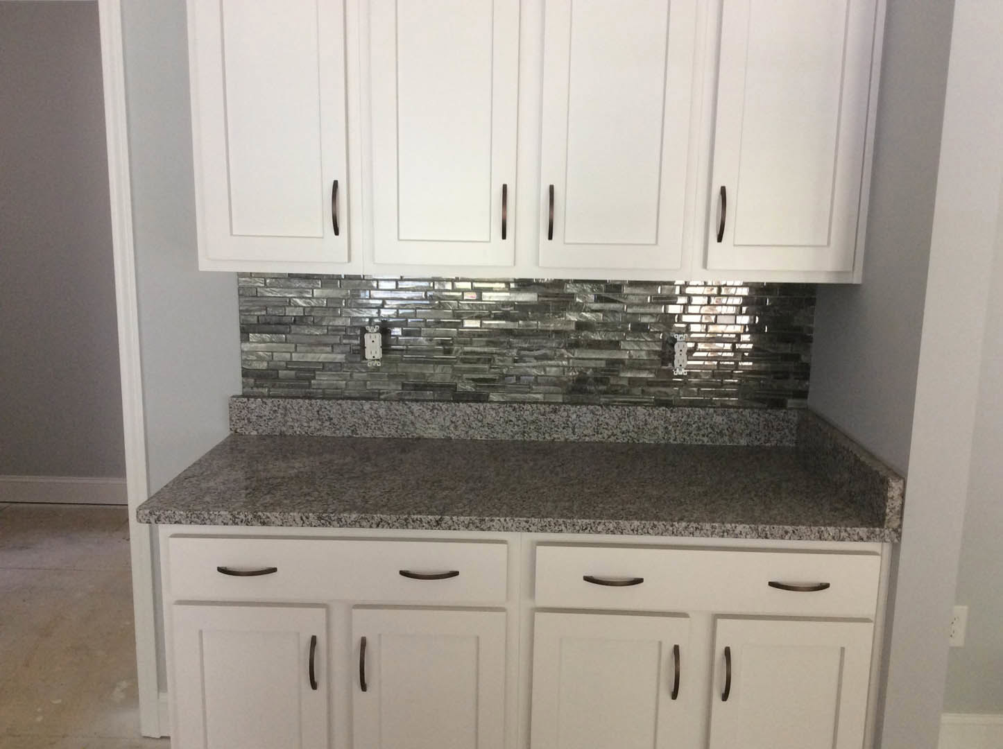 White kitchen cabinets with black handles, granite countertop, silver tile backsplash, and grey wall with white trim