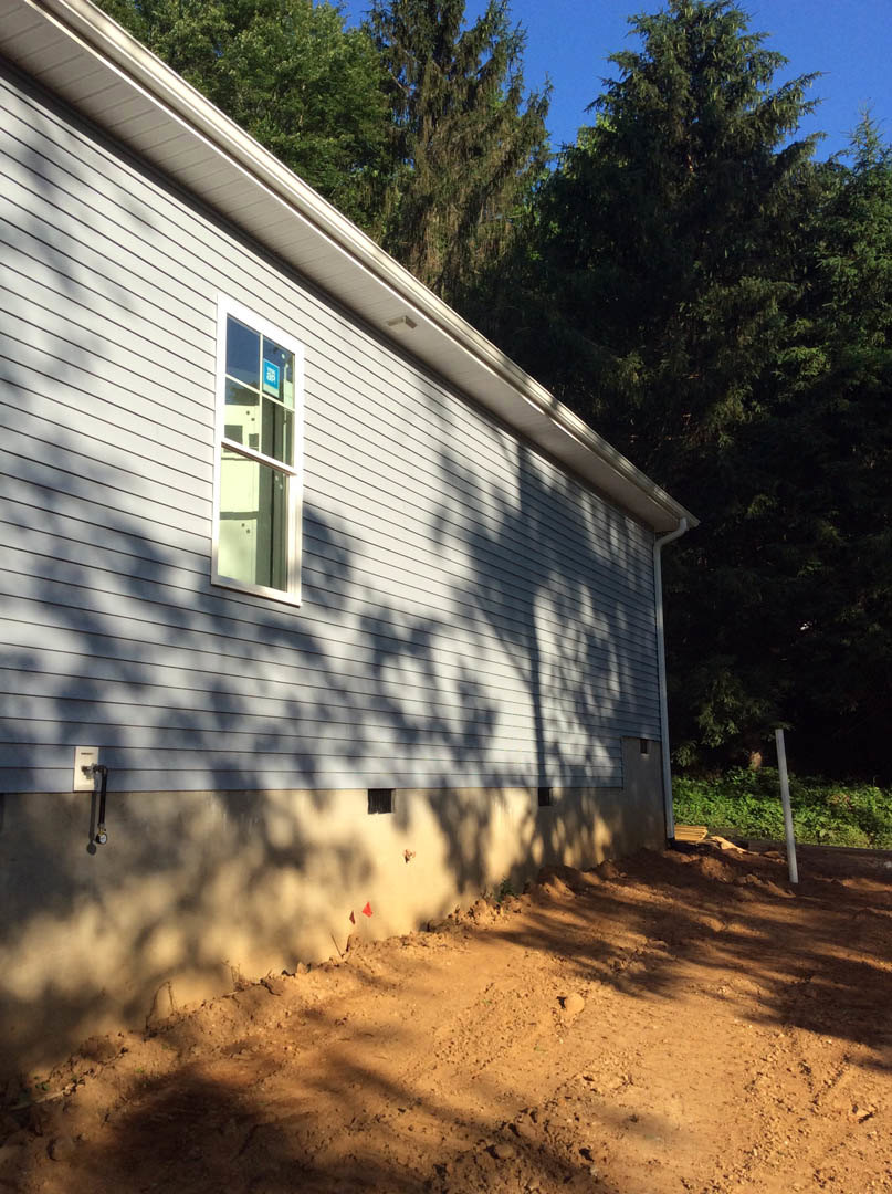 White house with large window and white frame, surrounded by mature trees, dirt road and bare ground in foreground, blue sky above
