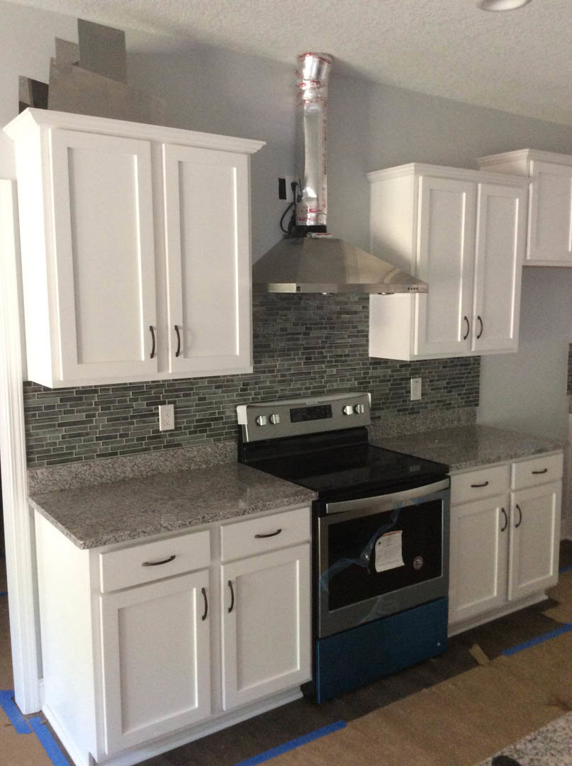 White kitchen with shaker cabinets, black handles, granite countertop, stainless steel stove, white curtain with red spots, paper resting on stove.
