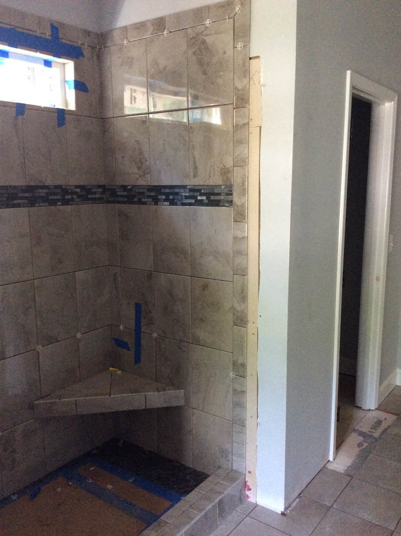 Bathroom corner with grey tile walls, marble flooring, a door with blue tape around the window, and a brick accent wall featuring a black stripe