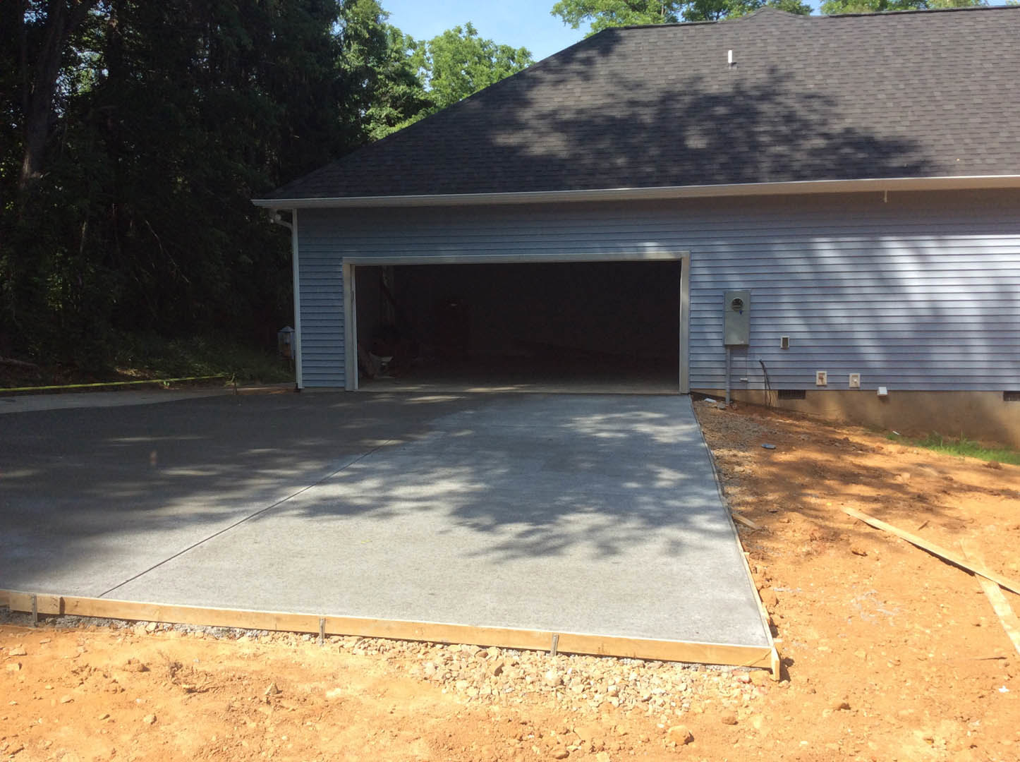 Garage with white paneled door, concrete driveway bordered by wood, shaded exterior with trees and blue sky in background