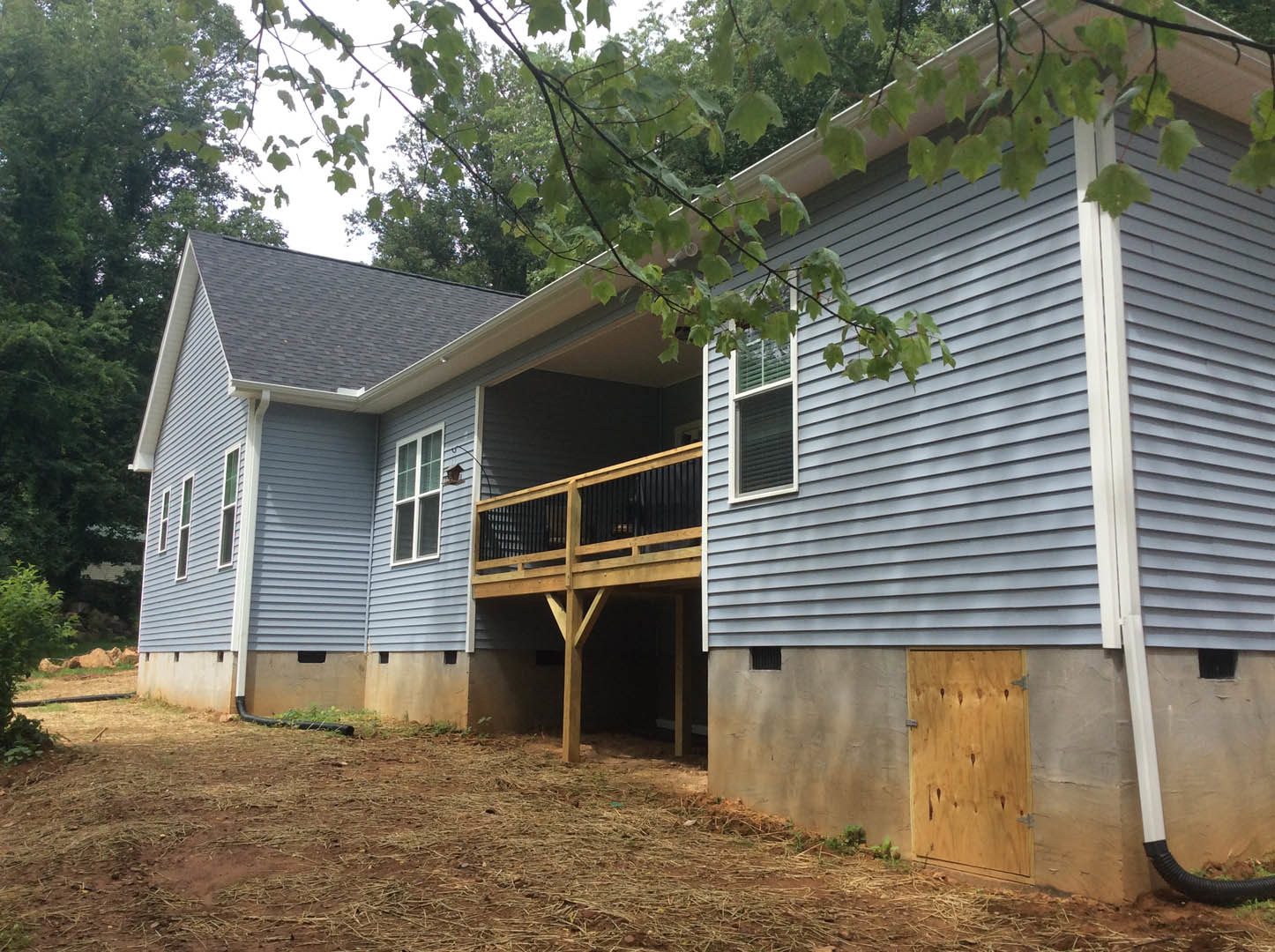 Wood siding cottage with open wooden door, metal hinges, covered porch, railing, window, leafy tree branches in foreground