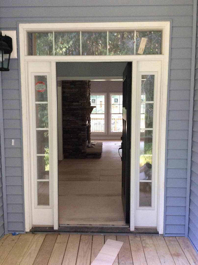 Wood front door with glass panels, stone porch floor, black accent chair, large window, and wood siding exterior