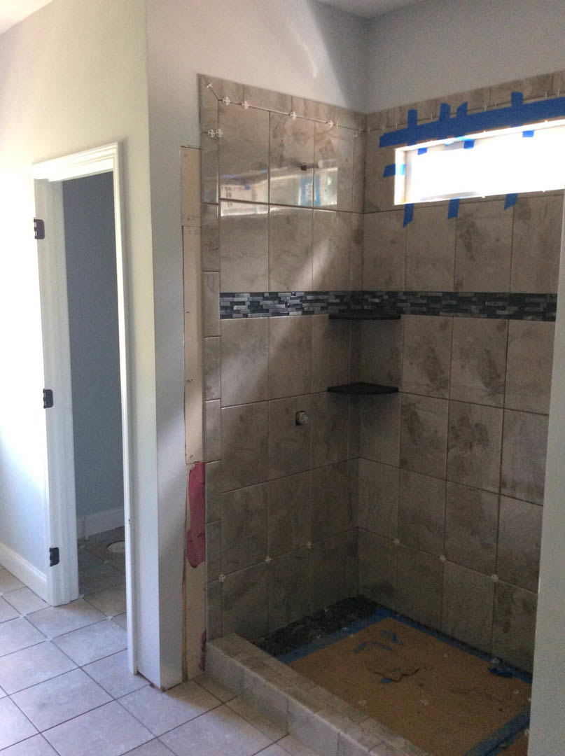 Bathroom with light gray tile walls, window with white trim, built-in shower shelf, blue painter’s tape on wall, and door partially visible.