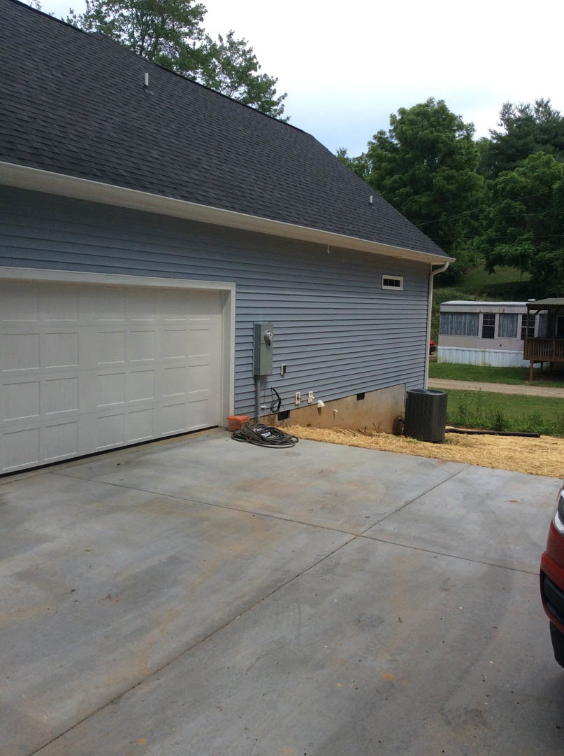 White garage door on a modern house, concrete driveway with black trash can and garden hose, windows and trees in background.