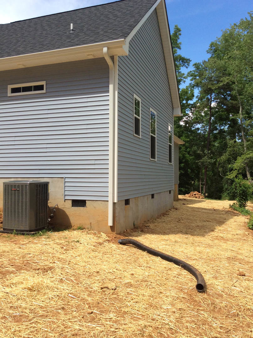 Modern home with light siding, large windows, and a leafy tree in front; long black pipe lies on hay-covered ground near a grey metal box.