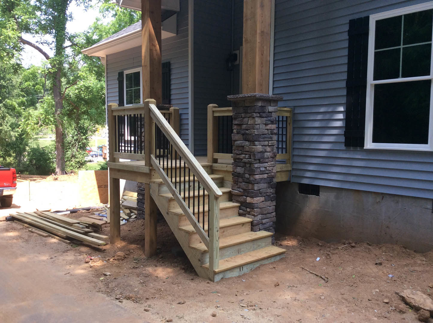 Wooden staircase with handrail leading to porch, stone pillar supporting column, close-up window, pile of lumber on ground, red truck parked near house, trees in background.