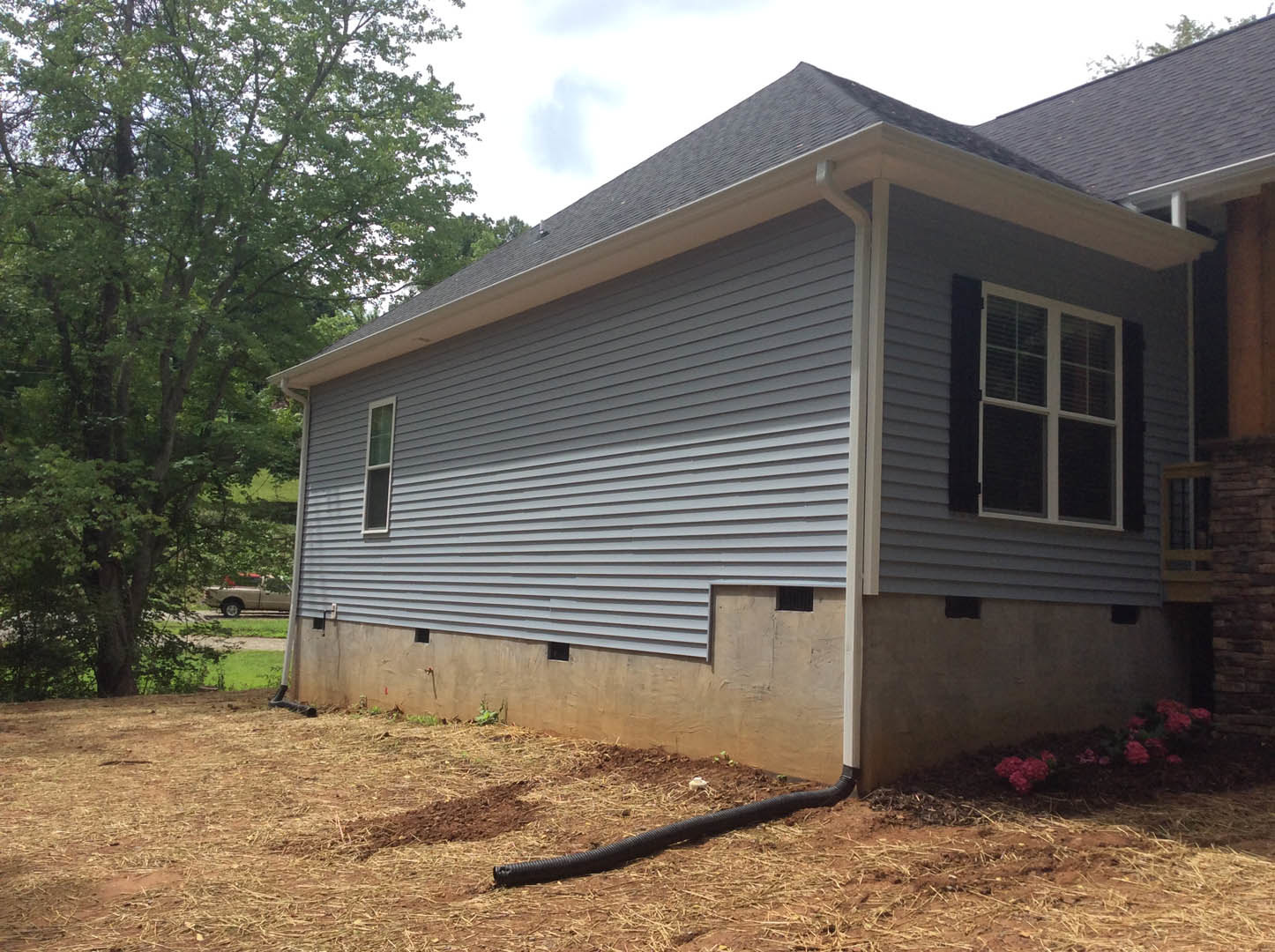 Grey siding house with white-framed window, black drain pipe along foundation, trees in background, partial view of a truck in foreground