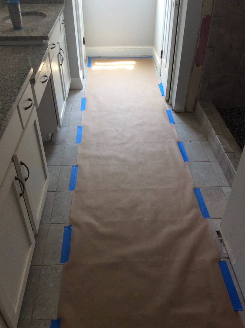Long carpeted hallway with strips of blue tape on the floor, white walls, recessed ceiling light, visible doorway, and a sink with a cup placed on the counter.