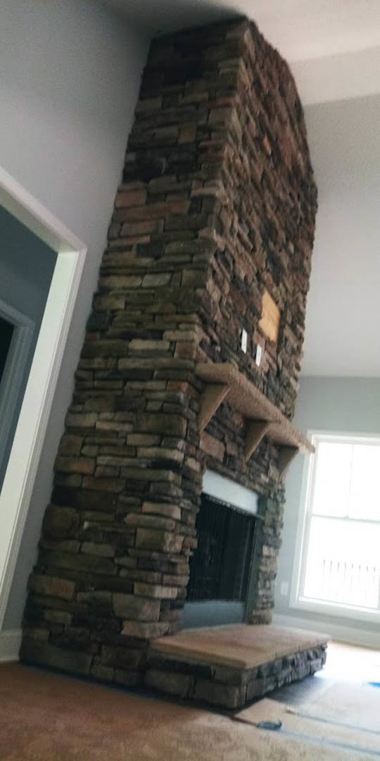 Stone fireplace with a built-in shelf, textured stone surround, adjacent window allowing natural light, hardwood flooring