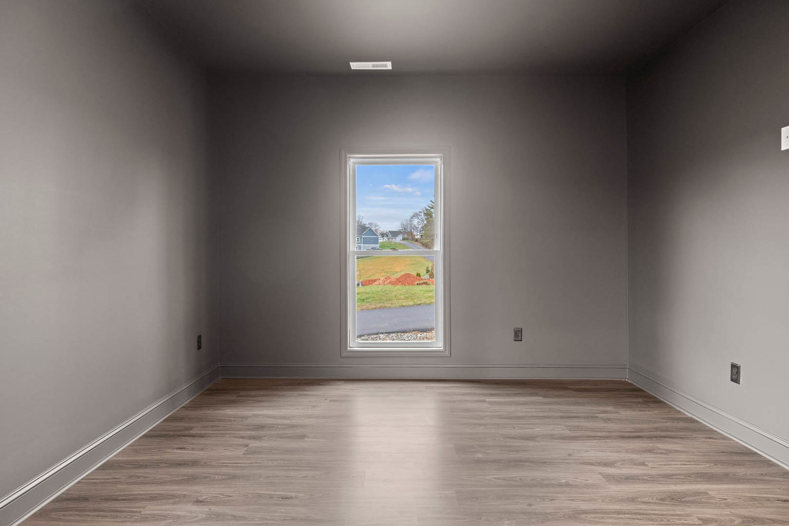 Sunlit room with wide window overlooking grassy field, smooth hardwood flooring, white plaster walls, and simple ceiling.
