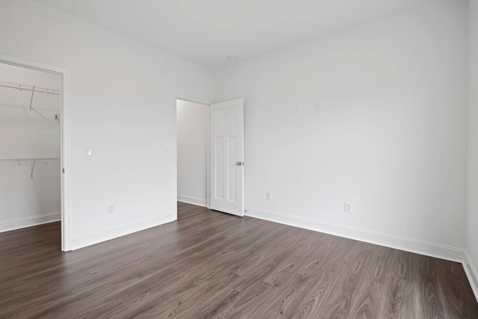 White paneled door with silver knob open onto wood laminate flooring, white walls, and white ceiling with fire sprinkler