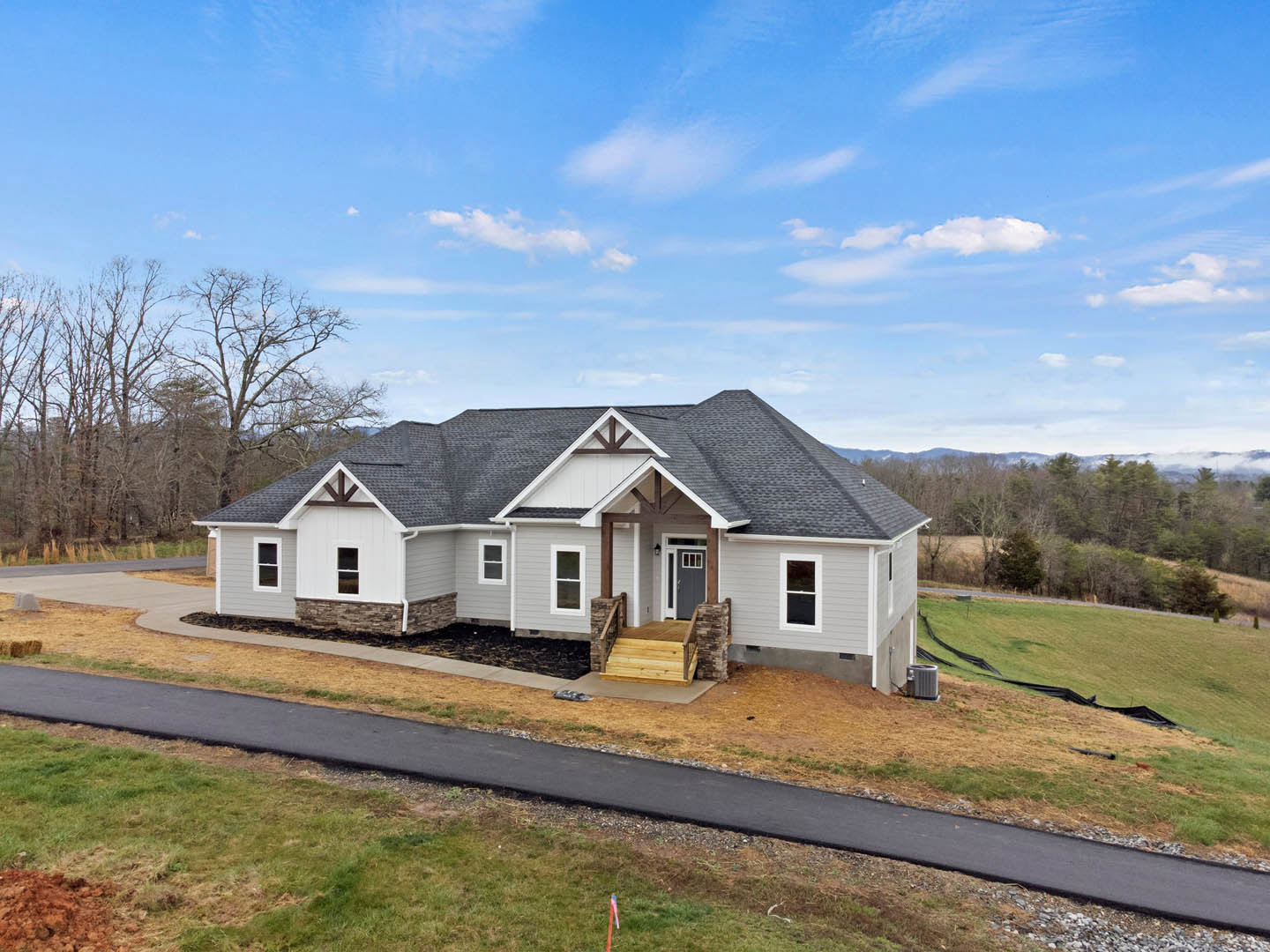 Two-story house with grey roof, wooden front steps, driveway bordered by green grass, trees in background, blue sky with scattered clouds