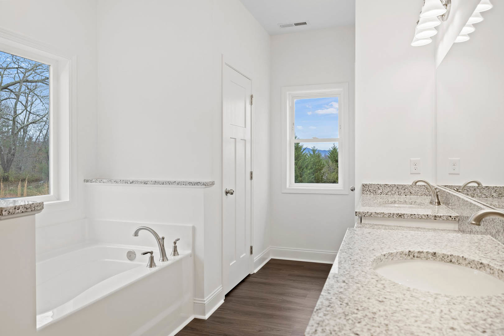 Bathroom with freestanding white bathtub, marble tile floors, white sink with black metal railing, row of recessed ceiling lights, window overlooking leafless tree, white door with