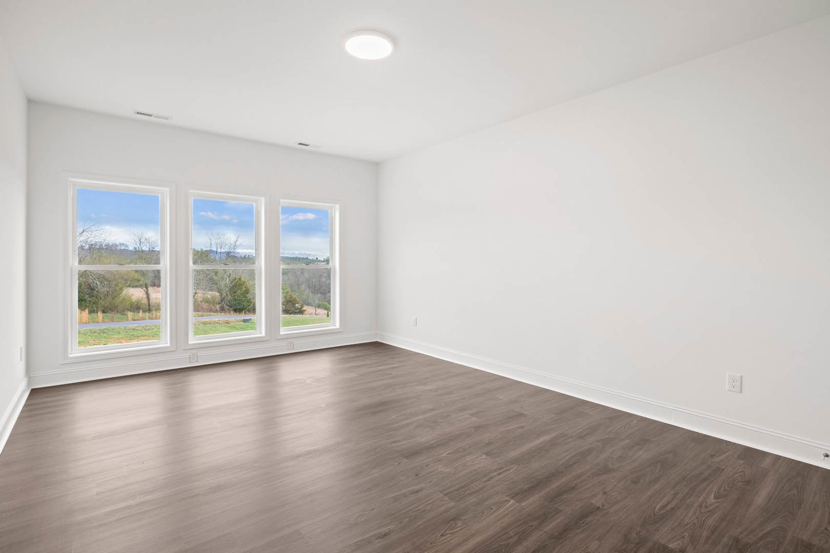 Spacious room with light wood flooring, large windows overlooking trees and grass, white ceiling with modern light fixture