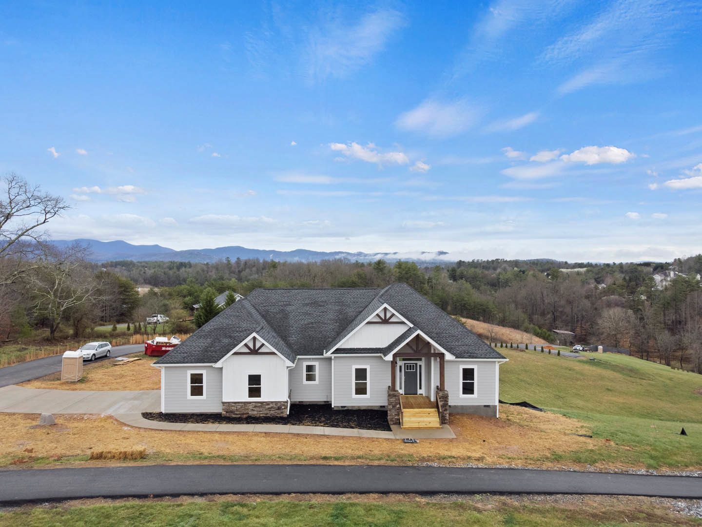 Two-story house with grey roof, white siding, and brown trim, surrounded by green grass and mature trees; concrete driveway leads to the front, blue sky with scattered clouds