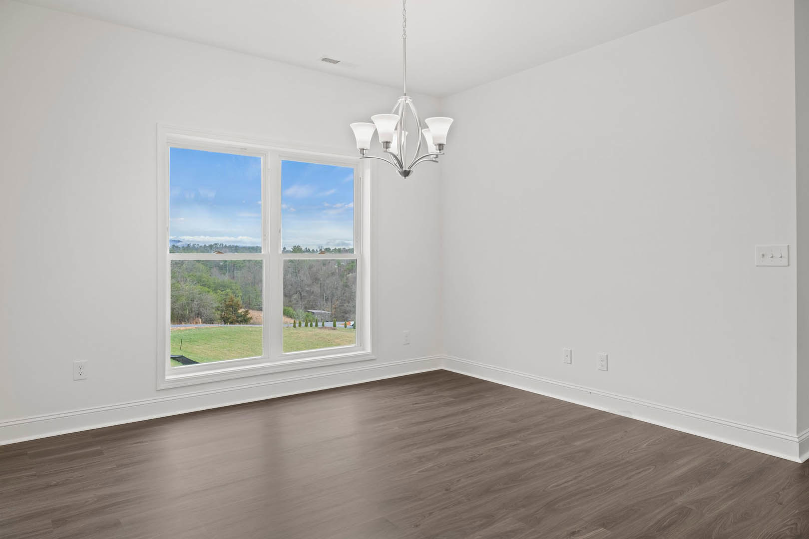 Bright room with wood flooring, large window overlooking green field and blue sky, white plaster walls, modern chandelier, and light switch visible
