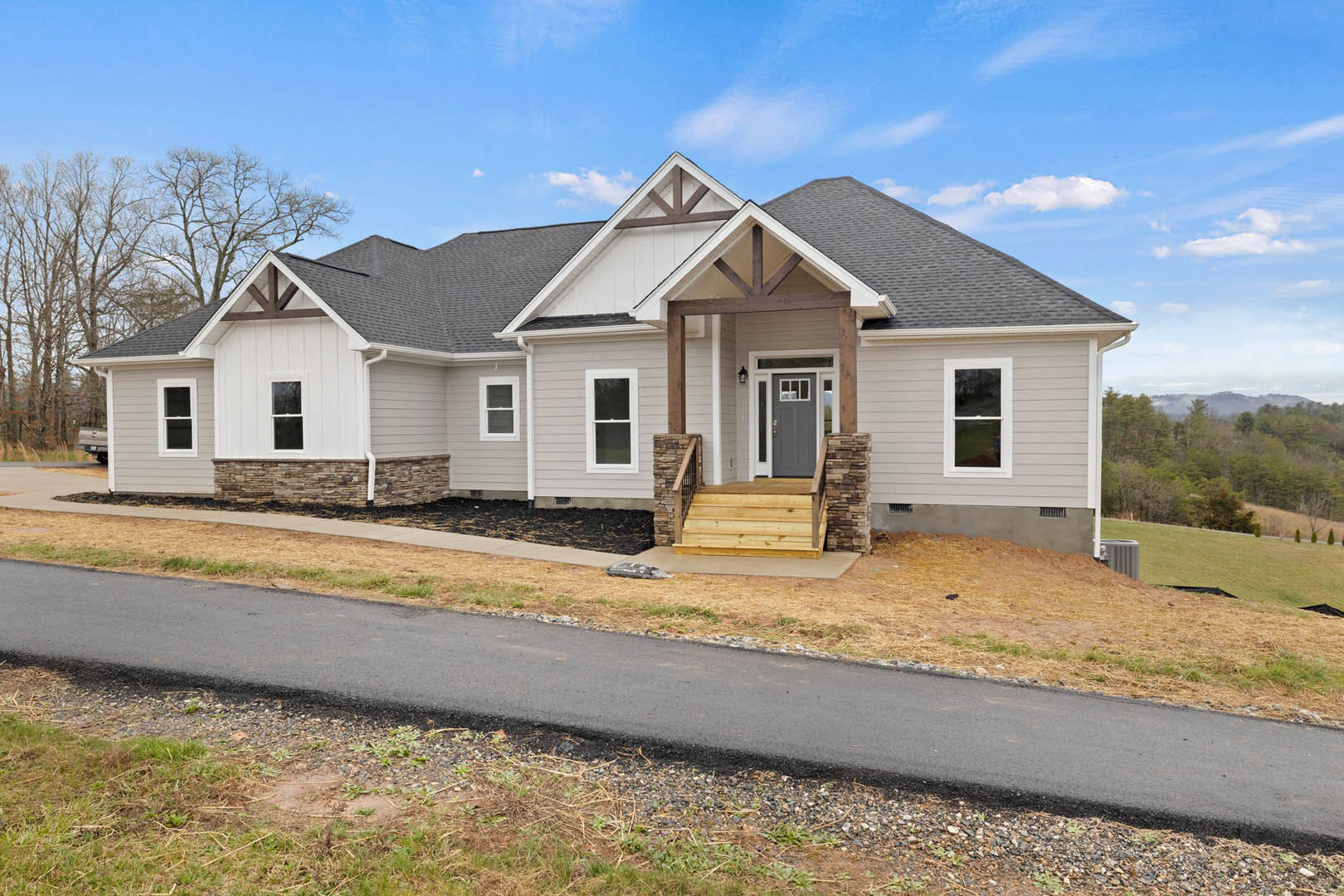 Two-story house with white-framed windows, wooden steps leading to front door, covered porch, gravel driveway bordered by grass, mature trees in background under partly cloudy sky